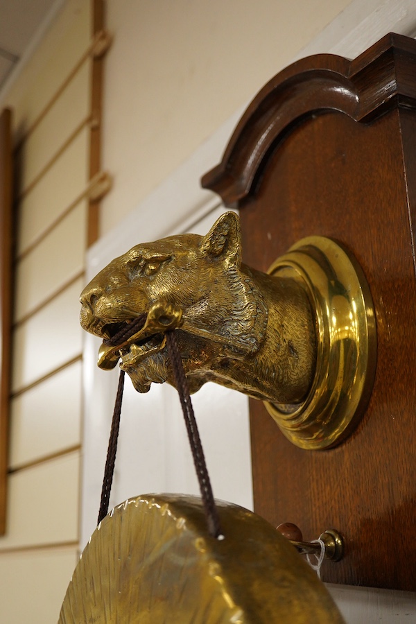 An early 20th century brass wall hanging gong, the holder in the form of a big cat, mounted on an oak plinth, the gong is supported on a bone clenched between its teeth, with the beater on a separate hook behind, 55cm to